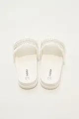 Sandalias de base plana color marfil con tira ancha decorada con bandas de cordón trenzado, detalles plateados con tachas y flecos en los bordes.