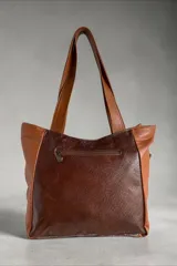 Cartera tipo tote de cuero en tono marrón, combinada con apliques de pelo vacuno animal print, detalles metalizados y dije colgante triangular.