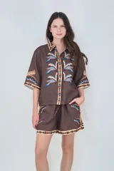 Camisa de lino marrón con estampado de hojas de palmera y detalles geométricos en color azul, celeste y naranja en el cuello, la tapeta y los puños. Tiene mangas cortas y corte recto.