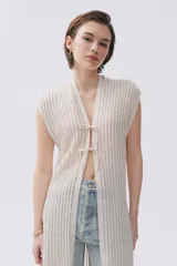 Kimono tejido de punto en color crudo con sutil brillo de lurex. Presenta un diseño sin mangas, corte largo y cierre frontal mediante dos lazos finos. El tejido tiene una textura acanalada vertical.