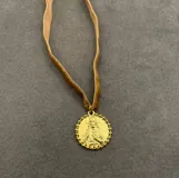 Collar tipo choker de cinta de terciopelo color tostado con dije circular dorado enchapado en oro 18k, con relieve de figura religiosa y borde texturado.