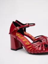 Sandalias de taco alto ancho con diseño retro, confeccionadas en charol negro. Presentan tiras delanteras múltiples entrelazadas, talón cerrado y una correa fina ajustable al tobillo con hebilla metálica.