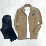 Sobrecamisa color beige de gabardina, con cuello clásico, cierre frontal con botones, dos bolsillos de parche con solapa abotonada en el pecho y parches bordados en el pecho y el hombro.