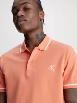Chomba color salmón de corte slim fit, con cuello clásico y tapeta de dos botones. Presenta ribetes blancos en cuello y mangas, y logo CK bordado en blanco en el pecho.