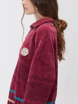 Pullover de punto con textura aterciopelada, diseño oversize, cuello polo con cierre frontal parcial y detalles de pespuntes en contraste. Presenta rayas horizontales en tonos tierra y un parche bordado con la letra M en el pecho.