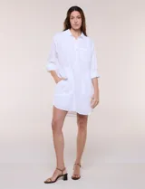 Vestido camisero corto de color blanco con sutiles rayas verticales, de la marca Dash. Presenta cuello con solapa, escote en V con botones parciales en el frente, bolsillo superior en el pecho y bolsillos laterales de gran tamaño. Cuenta con mangas largas de largo regulable mediante presilla y botón, calce holgado y ruedo trasero más largo.