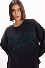 Buzo oversize de corte relajado con mangas amplias, cuello redondo y ruedo desflecado. Presenta letras bordadas en relieve con contorno en color verde azulado sobre fondo negro.