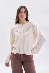 Blusa de estilo romántico en color crudo, confeccionada en tela texturizada con detalles de encaje y alforzas en el canesú. Presenta cuello redondo, cierre frontal con botones y mangas largas con puños delicados.