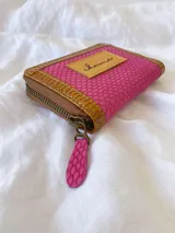 Billetera compacta de cuero con diseño bicolor en fucsia y marrón, con textura grabada tipo reptil. Cuenta con cierre metálico perimetral, tirador de cuero fucsia y etiqueta frontal con logo grabado.