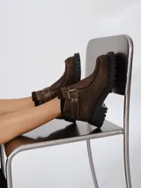 Bota corta tipo borcego de cuero negro con textura granulada, suela track dentada y tira decorativa con hebilla metálica en el tobillo.