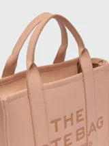 Cartera tipo tote de cuero granulado color tostado, con diseño estructurado y rectangular. Presenta doble asa superior fija y una correa larga ajustable y extraíble. En el frente, destaca el logo estampado con la inscripción 'THE TOTE BAG MARC JACOBS'.