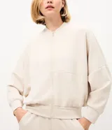 Campera bomber deportiva de mujer color beige, confeccionada en modal con cierre frontal por cremallera, cuello medio y bolsillos. Presenta mangas largas con puños elásticos y elástico en la cintura.
