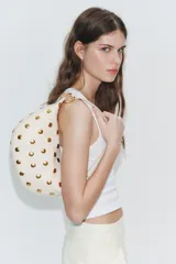 Bolso de hombro de diseño saca multiposición color blanco, decorado con tachas metálicas doradas y aros metálicos que conectan el cuerpo con la manija. Cuenta con cierre de cremallera metálica y bolsillo interior.