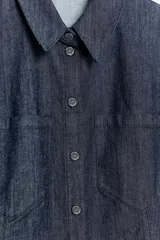 Camisa sin mangas de denim azul oscuro, con cuello solapa, hombreras marcadas, bolsillos delanteros de plastrón, bajo con peplum y cierre frontal con botones metálicos.