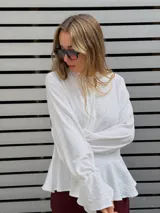 Blusa blanca de textura arrugada, con escote en V y lazos para anudar. Presenta mangas largas con puños acampanados y un diseño con volado en el ruedo.