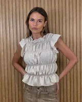 Blusa sin mangas de color beige, confeccionada con tejido texturizado. Presenta un diseño ajustado con múltiples costuras fruncidas mediante elásticos a lo largo de toda la prenda, formando volados sutiles en el cuello y en el ruedo.