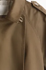Gabardina corta verde militar de doble botonadura, con solapas amplias, bolsillos laterales diagonales, charreteras en los hombros y mangas raglán amplias.