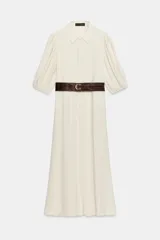 Vestido midi color crudo, con cuello camisero, mangas abullonadas por debajo del codo con puños elásticos y cierre frontal oculto con botones. Incluye bolsillos laterales y un cinturón ancho de cuero marrón oscuro con hebilla metálica.