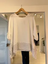 Blusa blanca de lino de corte oversize y asimétrico, con mangas tres cuartos y detalle de rayas negras en la parte inferior.