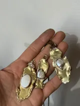 Collar artesanal con dije de base metálica irregular y piedra central roja. Presenta una estructura de tiento de gamuza marrón con detalles de tubos metálicos dorados.