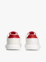 Zapatillas Tommy Hilfiger de diseño clásico y bajo, color blanco con detalles en rojo en la parte trasera del talón y en la lengüeta. Presentan cierre de cordones y el logo metálico distintivo de la marca en el lateral.