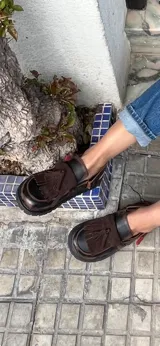 Mocasines de cuero color marrón oscuro, con diseño de flecos y tira con hebilla en el empeine, y suela plana con plataforma baja.