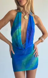 Vestido corto ajustado, sin mangas, con escote halter pronunciado y drapeado tipo cowl neck. Presenta un estampado abstracto en tonos azules y verdes que simulan olas o agua.