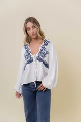 Blusa blanca de calce relajado con bordado frontal en azul de inspiración artesanal que realza el escote en V con lazo regulable. Posee mangas largas amplias con puño suave.