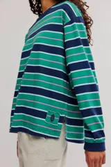 Remera manga larga de algodón con diseño de rayas horizontales en tonos verdes, azul marino, rosado y celeste. Presenta cuello redondo y un pequeño parche decorativo en el borde inferior.