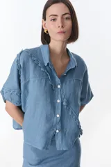 Camisa de lino color azul, de manga corta y cuello clásico. Presenta un diseño con botones frontales blancos y detalles de puntilla con ondas en diagonal sobre el frente y en las mangas.