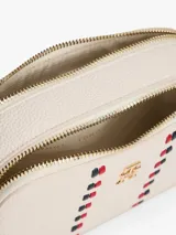 Cartera tipo bandolera de Tommy Hilfiger con acabado granulado, dos compartimentos principales con cierre de cremallera, correa ajustable y monograma TH metálico en la parte frontal junto a una cinta decorativa distintiva.