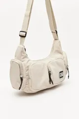 Cartera tipo shoulder color beige, de diseño utilitario y formato curvo. Confeccionada en material liviano y resistente, cuenta con correa regulable para llevar al hombro o cruzada. Presenta múltiples bolsillos con cierre en el frente y laterales, y un parche con el logo "Piece & Cake" en el frente.