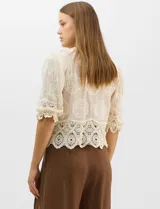 Blusa de algodón semitransparente con diseño floral bordado a tono. Presenta escote en V, cierre frontal con botones y mangas al codo ligeramente abullonadas. Incluye detalles de encaje calado estilo macramé en los puños y en el ruedo con terminaciones en ondas.