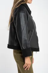 Campera tipo gamulán de cuero sintético negro con interior y detalles en corderito negro. Presenta cierre frontal, cuello solapa y diseño oversize.