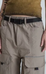 Bermuda técnica color arcilla (marrón claro/beige) de calce oversize, confeccionada en poliéster liviano. Presenta múltiples bolsillos cargo con tapa y un cinturón de ajuste con hebilla plástica negra. Incluye detalles reflectivos.