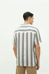 Camisa de manga corta con cuello cubano, confeccionada en una mezcla de lino y algodón con textura. Presenta un diseño a rayas verticales en tonos beige y celeste sobre fondo blanco, con cierre frontal de botones y logo bordado en el pecho.