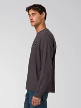 Remera de manga larga color gris oscuro, con cuello redondo y puños acanalados. Presenta un pequeño parche rectangular con el logo de la marca en el lado izquierdo del pecho.