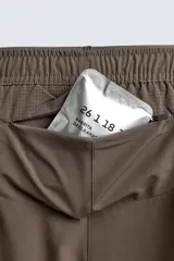Pantalón corto deportivo color marrón oliva, confeccionado en tejido técnico ligero y elástico, con forro interior. Presenta cintura elástica ajustable con cordón, bolsillos frontales, bolsillo de seguridad trasero con cierre y aberturas laterales en el bajo.