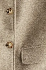 Campera corta de corte recto en color beige melange, con cuello camisero y cierre frontal mediante botones a contraste. Presenta dos bolsillos de parche con solapa en la parte delantera y cuenta con forro interior.