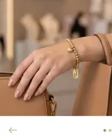 Pulsera elástica de acero dorado compuesta por pequeñas esferas, con un dije en forma de corazón y una placa rectangular con la inscripción 'Good Luck'.