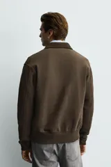Chaqueta de algodón con interior de rizo, color marrón con efecto lavado. Presenta cuello de pana, bolsillos frontales tipo canguro, puños y ruedo en rib, y cierre frontal con alamares metálicos.