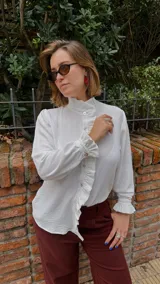 Camisa blanca de textura sutil, con cuello alto tipo victoriano y detalle de volados asimétricos a lo largo de la botonadura frontal. Presenta mangas largas con puños terminados en volados.
