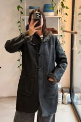 Campera de cuero genuino negro, estilo duffel coat con capucha, forro interno de corderito sintético, bolsillos frontales aplicados y cierre frontal con trabas de cuero y broches.