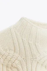 Chaleco de punto en color crudo con cuello subido y manga sisa. Presenta un diseño de tejido con estructura de cables gruesos en el frente y acabados acanalados en el cuello y el bajo.