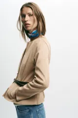 Chaqueta color beige con efecto ante, cuello redondo, manga larga y cierre frontal con cremallera. Presenta un bajo en evasé y un cinturón fino de cuero marrón oscuro con hebilla metálica.