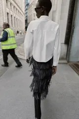 Camisa de popelín blanca con cuello solapa y manga larga. Presenta detalle de botones joya decorativos en forma de lazo en la parte frontal y cierre oculto por solapa.