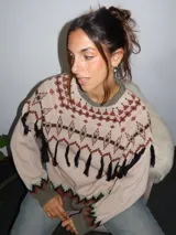 Sweater tejido con técnica intarsia, de cuello redondo, mangas abullonadas y silueta holgada con tajos laterales. Presenta un diseño geométrico con flecos decorativos en la parte frontal y detalles en puños y ruedo.