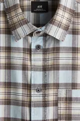 Camisa de manga larga con cuello inglés, tapeta clásica y canesú en la espalda. Presenta un bolsillo superior abierto y bajo redondeado. El tejido es suave de algodón con un patrón de cuadros en tonos celeste, marrón y beige.