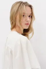 Blusa blanca de algodón con cuello redondo ribeteado en encaje y mangas cortas holgadas. Presenta un canesú fruncido con jaretas y detalles de broderie en la parte frontal.
