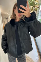 Chaqueta tipo gamulán de cuero genuino en color negro, con interior y cuello de piel sintética al tono. Presenta cierre frontal, diseño de paneles geométricos y corte holgado.